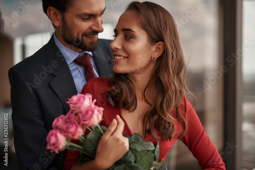 Concept of romantic date. Waist up portrait of happy beloved lady with pink roses looking tenderly to her male lover while standing in arms of each other
