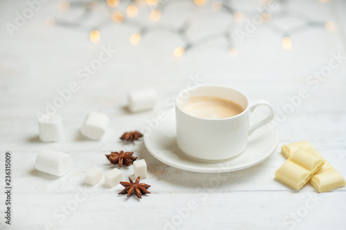Coffee with milk, latte with cinnamon sticks and anise stars with white chocolate and marshmallow, on a light white background, with lights from a garland.