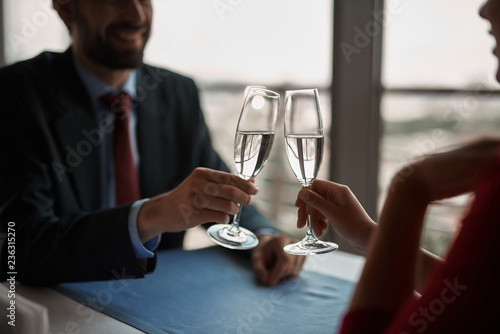 Concept of romantic date. Close up of two clinking glasses with champagne in hands of beloved couple sitting in restaurant