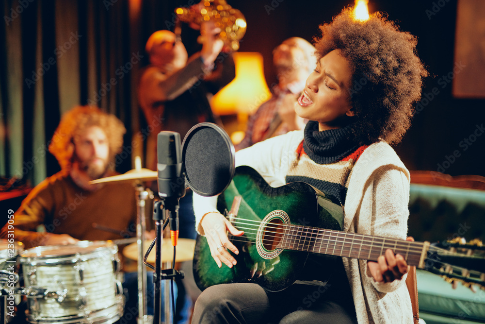 Fototapeta premium Mixed race woman singing and playing guitar while sitting on chair with legs crossed. In background drummer, saxopxonist and bass guitarist.