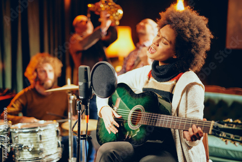 Mixed race woman singing and playing guitar while sitting on chair with legs crossed. In background drummer, saxopxonist and bass guitarist.
