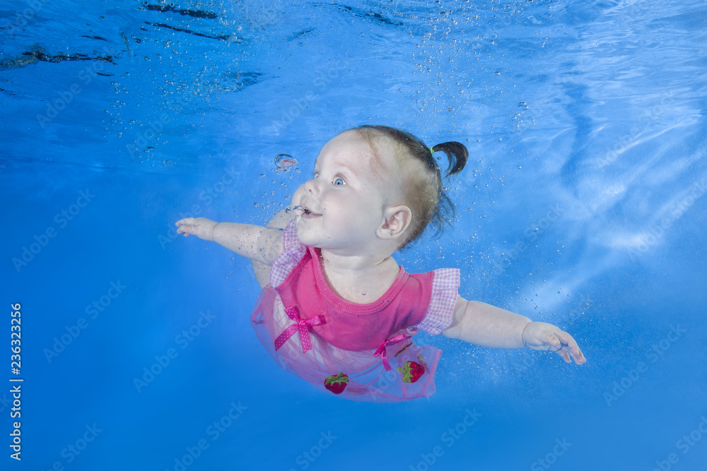 Little baby in pink dress swim underwater. Baby swimming underwater in the pool on a blue water background. Healthy family lifestyle and children water sports activity.