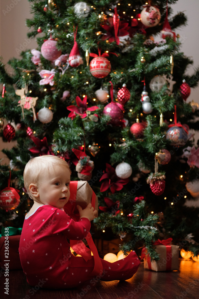 Little baby in red pajamas on the background of the Christmas tree with a gift in the festive package. Little baby in red pajamas on the background of the Christmas tree with a gift in the festive package.