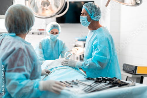 Side view portrait of focused doctor in sterile gloves holding laparoscopic instrument while looking away with serious expression. Nurses checking patient condition on blurred background