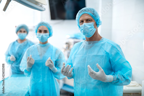 Best team. Waist up portrait of surgeon in protective mask and sterile gloves looking at camera with serious expression while his assistants standing behind him on blurred background photo