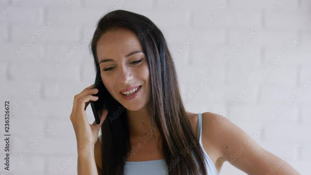 Beautiful young lady with brown hair smiling and talking on modern smartphone while standing on background of white wall Beautiful young lady with brown hair smiling and talking on modern smartphone while standing on background of white wall
