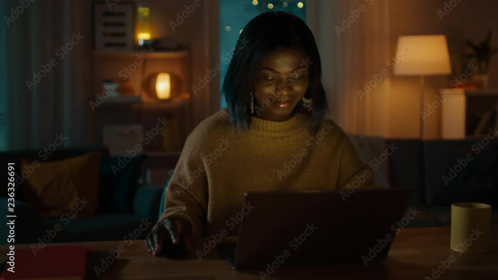 Portrait of Beautiful Smiling African American Girl Using Laptop while Sitting at Her Desk at Home. In the Evening Creative Woman Works on a Computer In Her Cozy Living Room. Portrait of Beautiful Smiling African American Girl Using Laptop while Sitting at Her Desk at Home. In the Evening Creative Woman Works on a Computer In Her Cozy Living Room.