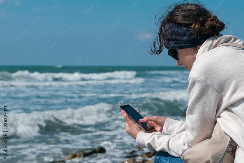 Woman is typing a message on phone sitting on the sea shore in autumn.