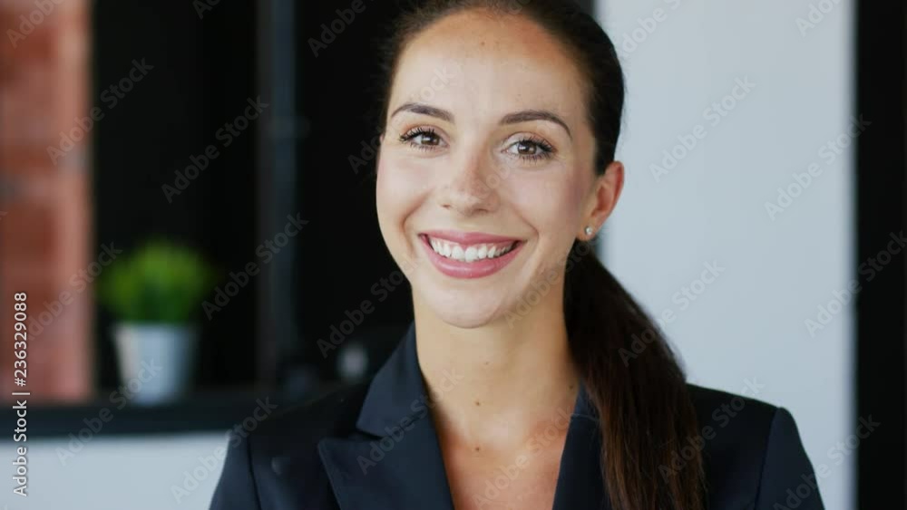 Confident young brunette in black jacket smiling and looking at camera while standing on blurred background of stylish room Confident young brunette in black jacket smiling and looking at camera while standing on blurred background of stylish room