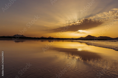 Famara Beach, Lanzarote, Spain. Orange sunset over the sea with clouds reflected in the wet sand of a large beach bordered by distant mountain silhouettes.