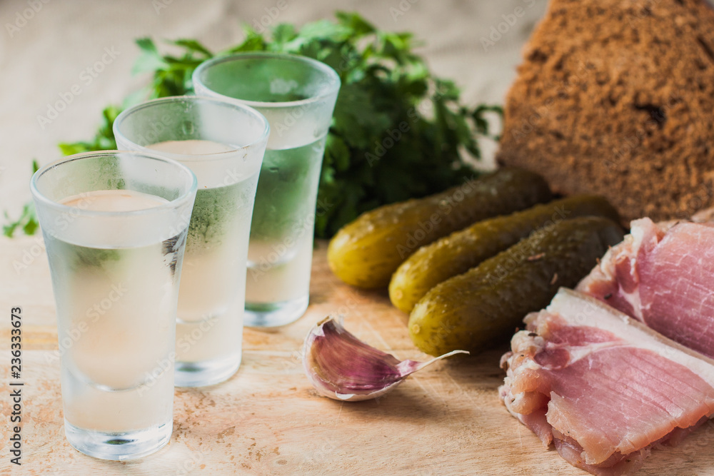 Φωτογραφία A shot of vodka on a blurry snack background - slices of bacon, garlic, cucumber