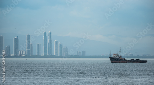 Large tug boat near the Panama Canal sits anchored and idle in hot humid weather. Panama City shimmers in rising heat waves as a summer storm approaches it from behind.