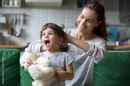 Smiling young loving mum making ponytail to little preschool laughing daughter with toy sitting on sofa, couch in living room at home, mother helping child with hairstyle, nanny caring of pupil