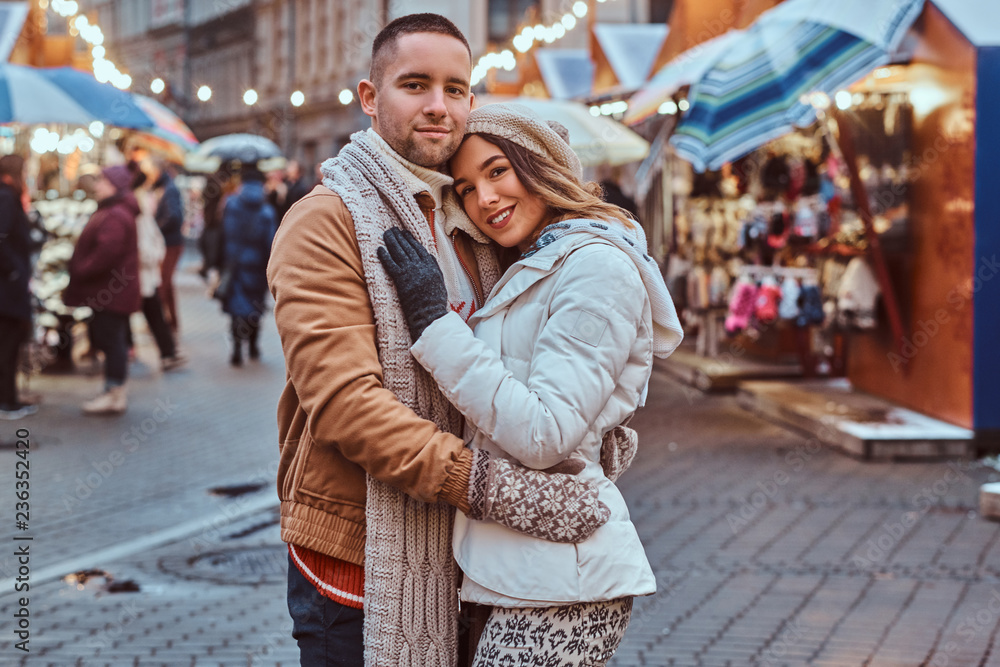 A young romantic couple wearing warm clothes hugging outdoor in evening street at Christmas time, enjoying spending time together.