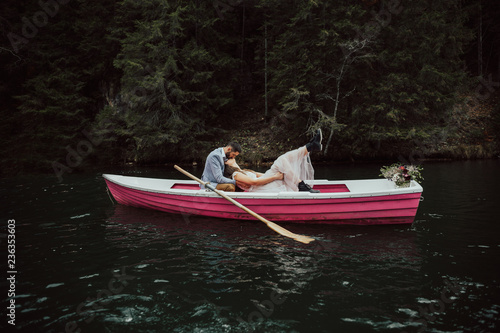 Wedding kiss on the retro boat.Bride and groom sitting in pink boat floating on the lake.Beautiful newly married couple kissing on rowing boat in the middle of lake.Nearby is a rustic style bouquet.