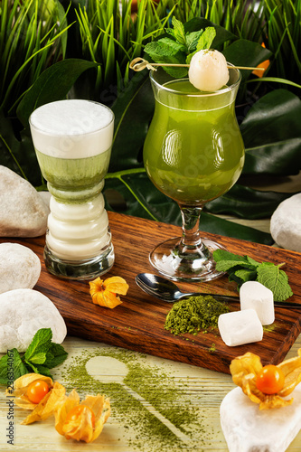 Glass of matcha green tea decorated with milk foam and marshmallow on a wooden board with tea powder at decorated background. Concept of healthy lifestyle