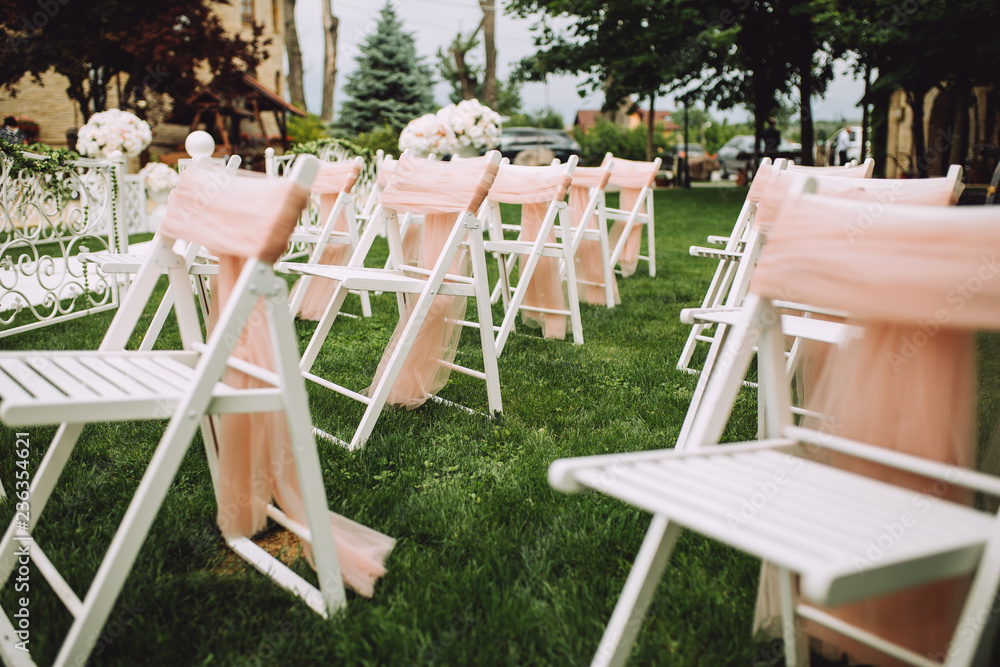 Rows of white empty chairs on a lawn before a wedding ceremony decorated by pink ribbons.Nobody in the place. 