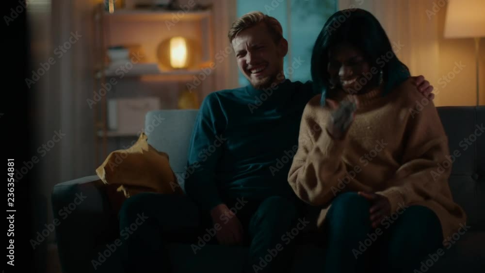 Happy Diverse Young Couple Watching Comedy on TV while Sitting on a Couch, they Laugh and Enjoy Show. Handsome Caucasian Boy and Black Girl in Love Spending Time Together. Moving Camera Shot. Happy Diverse Young Couple Watching Comedy on TV while Sitting on a Couch, they Laugh and Enjoy Show. Handsome Caucasian Boy and Black Girl in Love Spending Time Together. Moving Camera Shot.