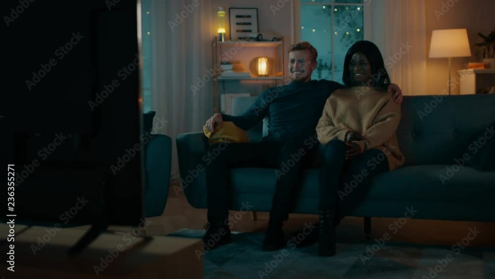 Happy Diverse Young Couple Watching Movie on TV while Sitting on a Couch, they Laugh and Enjoy Show. Handsome Caucasian Boy and Black Girl in Love Spending Time Together. Moving Camera Shot. Happy Diverse Young Couple Watching Movie on TV while Sitting on a Couch, they Laugh and Enjoy Show. Handsome Caucasian Boy and Black Girl in Love Spending Time Together. Moving Camera Shot.