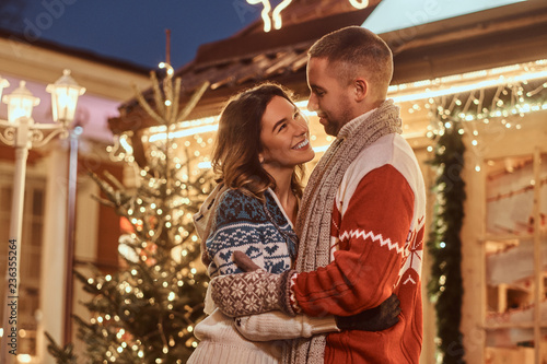 A young romantic couple wearing warm clothes hugging outdoor at Christmas time, standing in evening street decorated with beautiful lights.