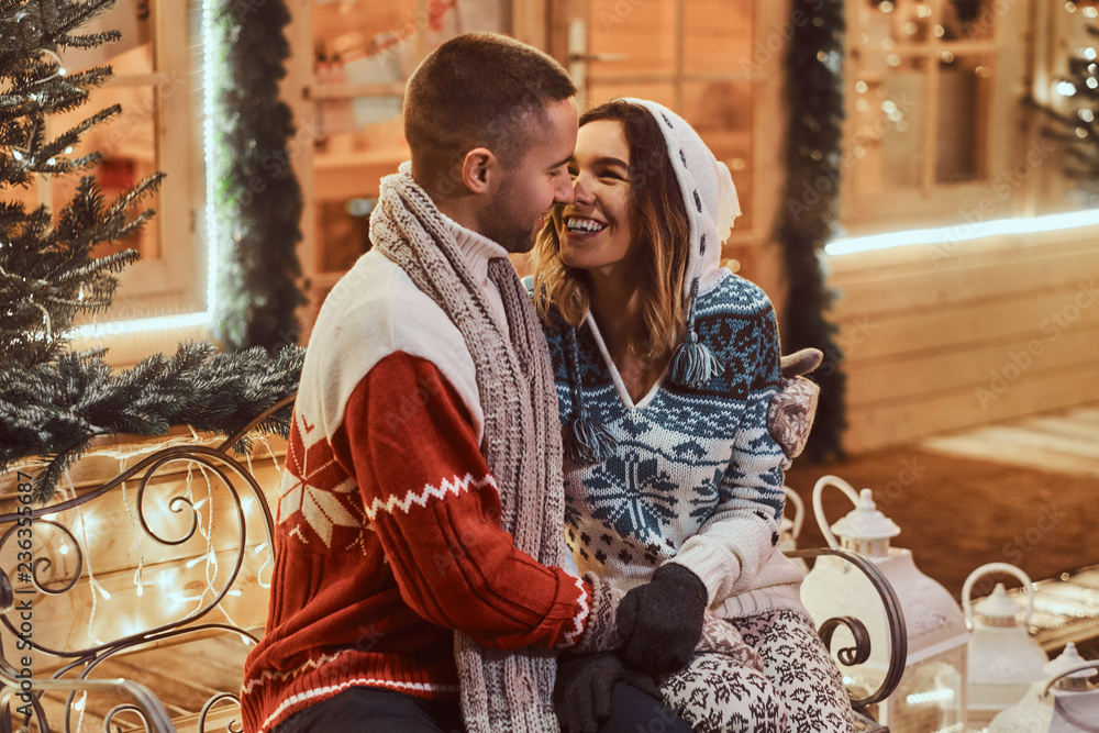 A young romantic couple wearing warm clothes outdoor at Christmas time, sitting on a bench in evening street decorated with beautiful lights.
