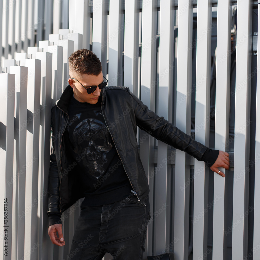 Fashionable handsome young model man with sunglasses in black leather jacket  with black pullover poses near the metal wall Stock-Foto | Adobe Stock