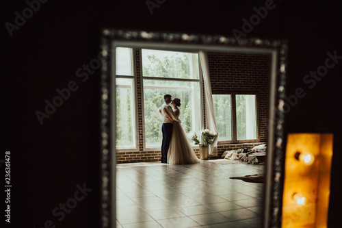 Young man gently embracing his bride and looking with love into her eyes.Newlyweds at wedding day.Happy luxury bride and groom standing at window light in rich room, tender moment.