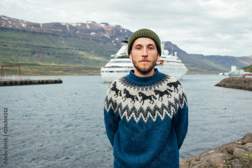 Portrait of young man, future sailor or captain in fisherman outfit, green beanie hat and blue traditional wool sweater with authentic ornament stand in front of big cruise ship or boat entering port