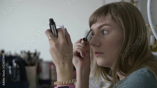 Young beautiful blonde woman applying mascara for eyebrows on her face doing professional trendy makeup indoors.