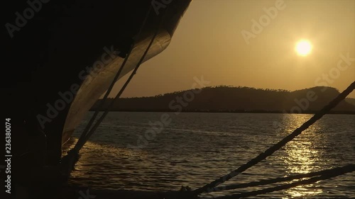 wide still perfect full, sunset view shot, bright ball of yellow-gold rays, clear light orange sky, reflection on grey quiet Conakry port water, ship's brown bow, thick hanging docking ropes, sundown