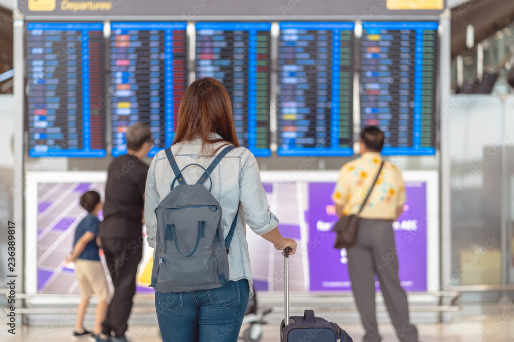 Back side of asian woman traveler with luggage standing over the flight board for check-in at the flight information screen in modern an airport, travel and transportation concept