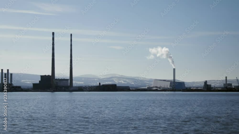 Factory in winter on water in Dublin Ireland - Static shot of Dublin factories in operation as water passes by in foreground. Snowy Irish hills visible in the distance Factory in winter on water in Dublin Ireland - Static shot of Dublin factories in operation as water passes by in foreground. Snowy Irish hills visible in the distance
