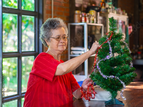Senior woman decorating the christmas tree with white and red ribbons at home.