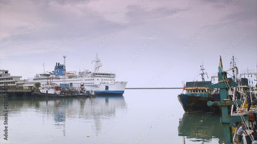 Wide still side view shot of large Africa Mercy ship, tugboat, at docking terminal, Conakry; front of old, rusty vessels, parts secured by ropes; radar masts, floodlight structures point light cloudy Wide still side view shot of large Africa Mercy ship, tugboat, at docking terminal, Conakry; front of old, rusty vessels, parts secured by ropes; radar masts, floodlight structures point light cloudy