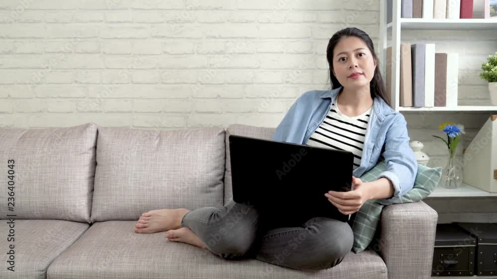 housewife using laptop shopping online store on summer sale and holding controller switching tv. happy asian lady watching funny news and laughing relaxing on couch in living room in daytime alone. housewife using laptop shopping online store on summer sale and holding controller switching tv. happy asian lady watching funny news and laughing relaxing on couch in living room in daytime alone.