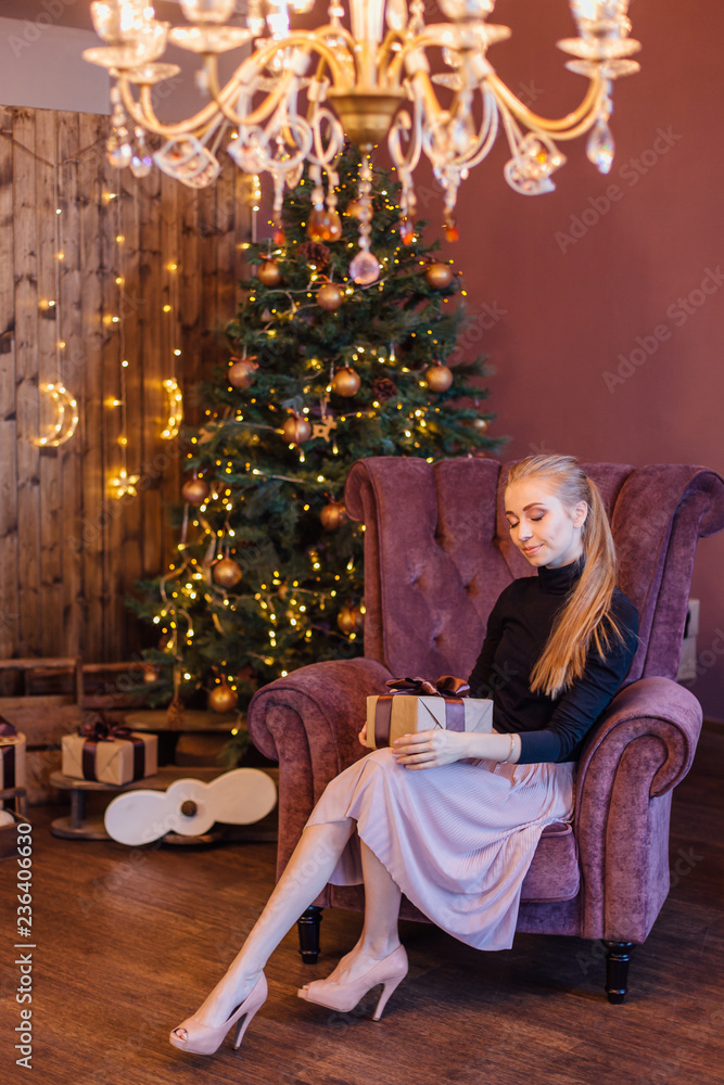 Beautiful young woman dressed in a sweater and skirt sitting on the arm chair next to the christmas tree with gift box in hands.