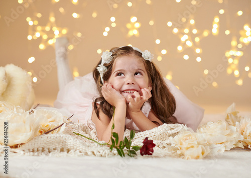 girl child with rose flower is posing in christmas lights, yellow background, pink dress