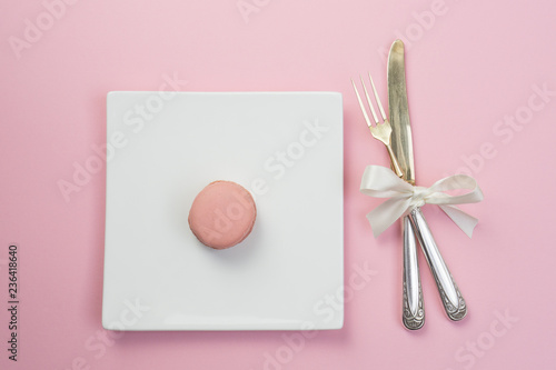 Silver fork and knife with white ribbon and plate with a maracon on pink background