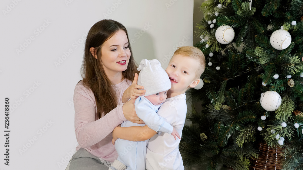 Young woman and boy embracing cute baby while standing near beautifully decorated Christmas tree at home Young woman and boy embracing cute baby while standing near beautifully decorated Christmas tree at home
