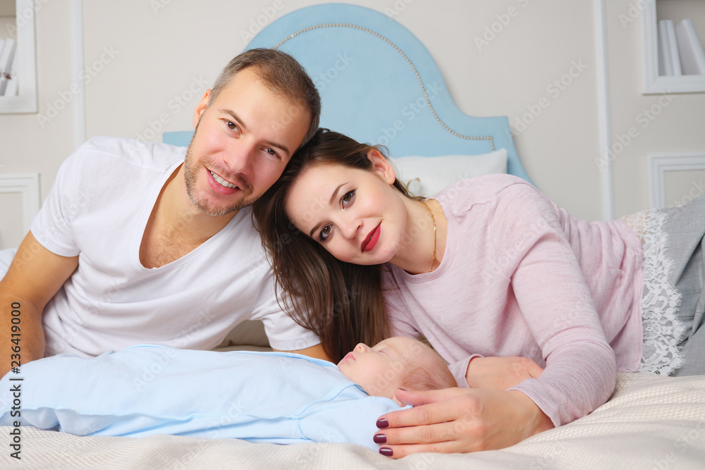 Happy parents while lying on soft bed near sweet sleeping baby in stylish bedroom, looking at camera - obrazy, fototapety, plakaty 