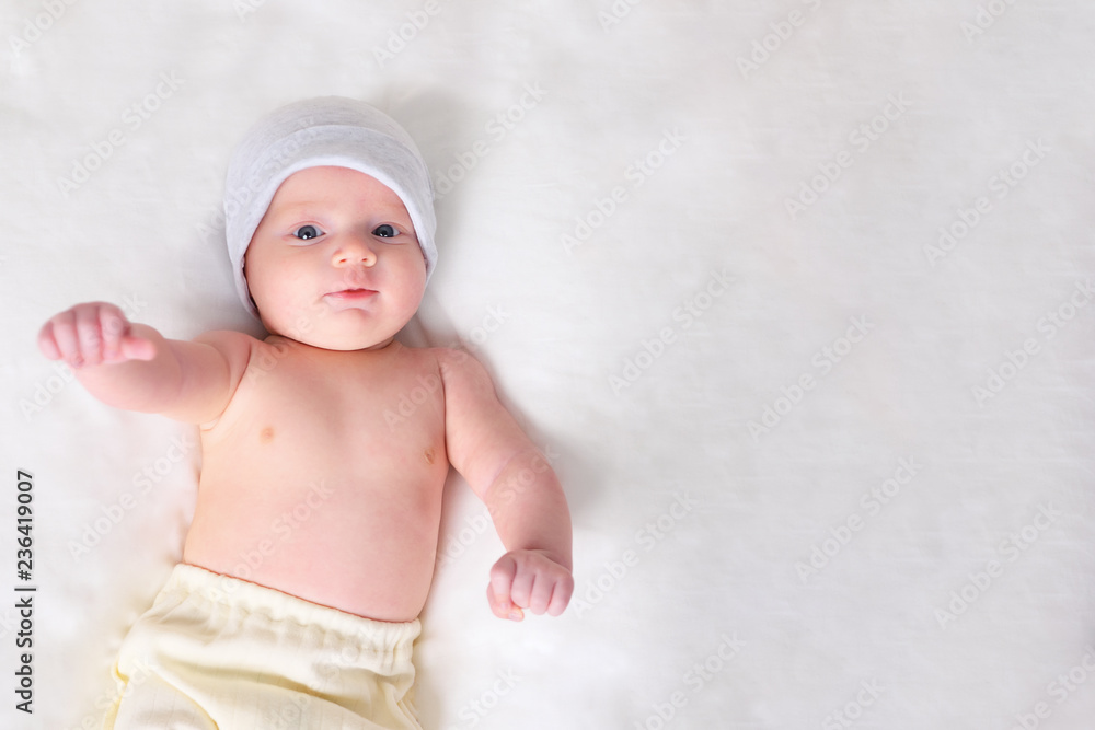 From above shot of sweet baby in hat and pants looking at camera while lying on grey background. From above shot of sweet baby in hat and pants looking at camera while lying on grey background.