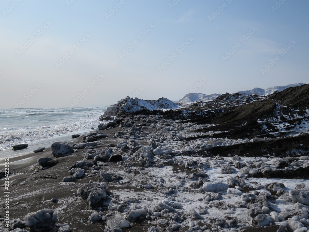 Pacific Ocean, waves and views of the snow-covered hill in winter in sunny weather in Kamchatka, Russia