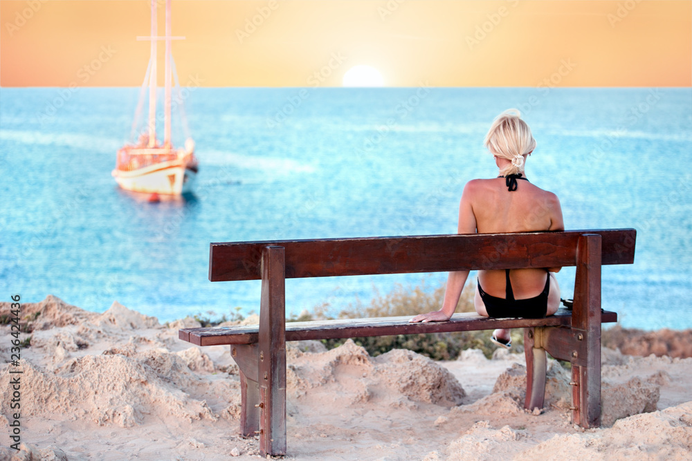 woman in swimsuit sitting on the bench looking sea, sunset, sailboat. relax travel concept.