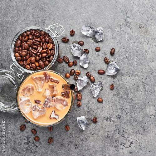 Cold brewed iced coffee in glass and coffee beans in glass jar on grey concrete background. Chocolate, vanilla, caramel or cinnamon iced coffee in tall glass. Top view, square crop.