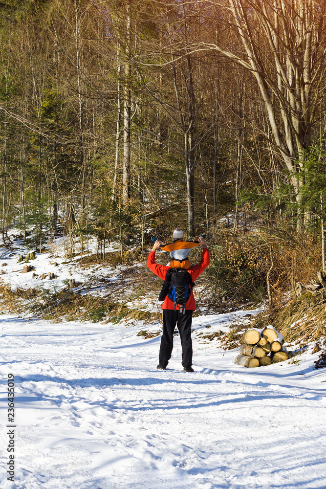 Man with backpack and son on shoulders stands with arms raised against the background of coniferous trees in the forest. Winter day. View from the back. Vertical frame