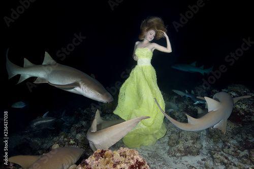 Night dance with sharks. Young beautiful woman in yellow dress dancing underwater with Tawny Nurse Sharks (Nebrius ferrugineus), night shooting in Indian Ocean on Maldives photo