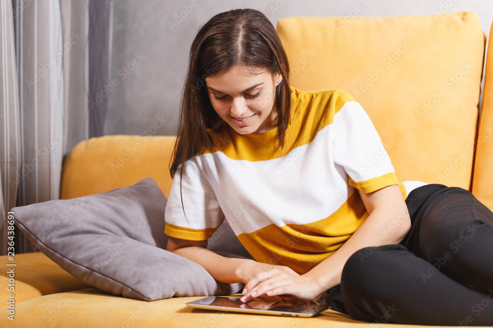 Cute long haired brunette girl laying on the yellow sofa in the light living room and searching something on the digital tablet, modern technology, communication concept Cute long haired brunette girl laying on the yellow sofa in the light living room and searching something on the digital tablet, modern technology, communication concept