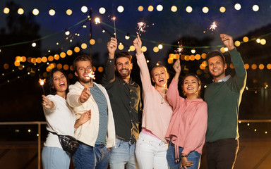 leisure, celebration and people concept - happy friends with sparklers at rooftop party at night with blurred bokeh lights