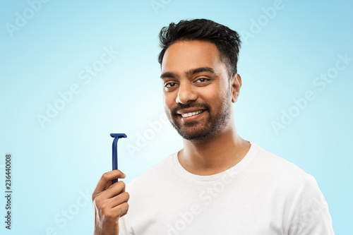 beard shaving, grooming and people concept - smiling indian man with manual razor blade over blue background