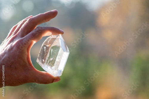A Hand holding a transparent crystal quartz with the sun hitting the gemstone and a blurry background photo